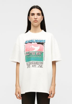 Kvinde med langt mørkt hår iført en oversized hvid T-shirt med farverig grafisk tekst og design foran, stående mod en ensfarvet baggrund.