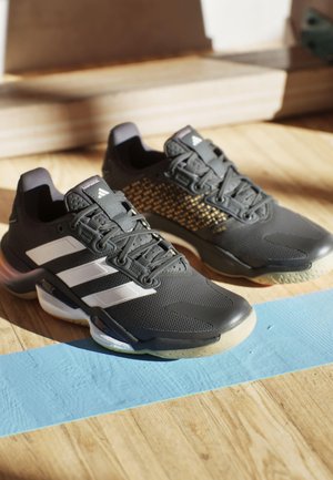 Scarpe da ginnastica nere con strisce bianche, parte superiore strutturata e un motivo in oro. Suola in gomma con elementi trasparenti.