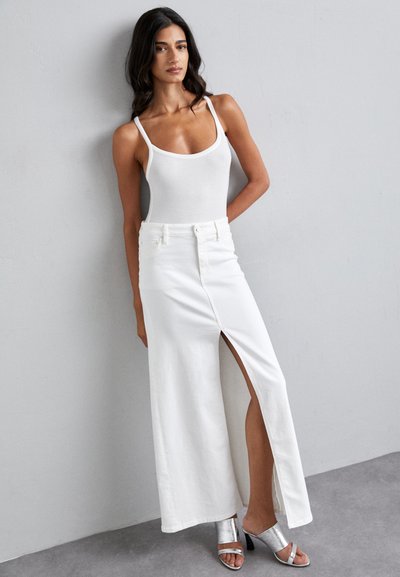 Falda maxi de algodón blanca con una abertura alta, combinada con una camiseta ajustada blanca y sandalias de tacón plateadas, sobre un fondo neutro.