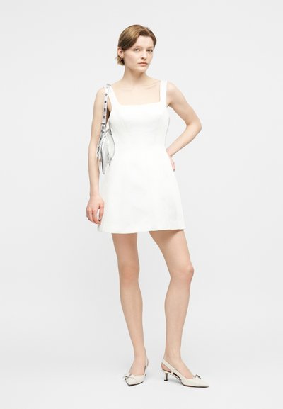 Femme portant une mini robe blanche sans manches, des chaussures blanches à talons bas et un sac à bandoulière argenté avec des franges, debout devant un fond uni.