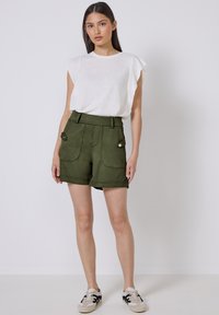 Bílý top s krátkým rukávem spárovaný se zelenými šortkami s dvěma předními kapsami a knoflíkovými detaily. Modelka v teniskách stojí před jednolitým pozadím.