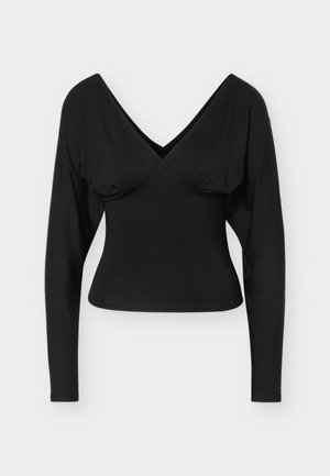 Melnas garām piedurknēm pieguļošs tops ar dziļu V veida izgriezumu un nelielu savilkumu zem krūtīm, uz gaiša, vienkārša fona.