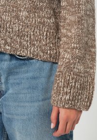 Kötött pulóver texturált, világosbarna és krémszínű mintával, bordázott mandzsettával. Világoskék farmernadrággal párosítva.