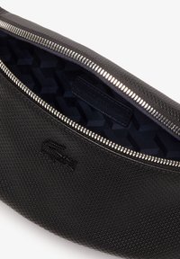 Pochette noire texturée avec fermeture éclair argentée partiellement ouverte, dévoilant un tissu intérieur sombre à motif et un logo crocodile embossé à l'extérieur.