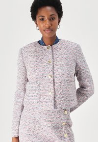 Veste en tweed avec un col rond, tissu texturé rose et bleu, et boutons en forme de cœur dorés. Elle possède des manches longues et une coupe ajustée.