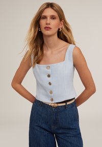 Ljusblå topp med fyrkantig halsringning gjord av texturerat tyg, med fem knappar fram och ihop med mörkblå högmidjade jeans.