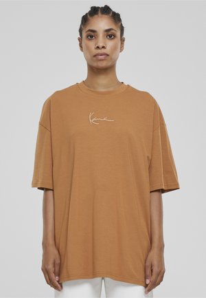 Kobieta w luźnej, brązowej koszulce z krótkim rękawem, z białym, kursywnym napisem na piersi, stojąca na jednolitym, jasnym tle.