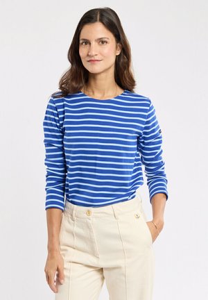 Femme aux cheveux foncés mi-longs portant une chemise à manches longues rayée bleu et blanc et un pantalon crème taille haute, debout avec une main dans la poche.