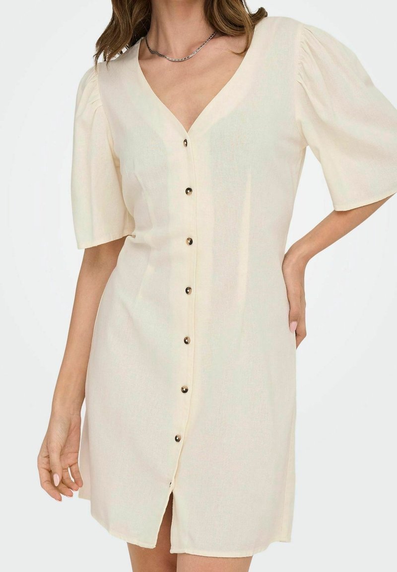 Femme portant une robe boutonnée crème à manches courtes bouffantes et col en V, debout avec une main sur la hanche.