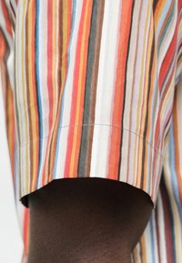 Chemise en coton présentant des rayures verticales rouges, oranges, jaunes, brunes et blanches. Manches coupées avec un ourlet cousu. Texture lisse.