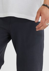 Muž v tmavých kalhotách a bílém tričku, ukazující ruku s několika náramky a prsteny, stojící před jednoduchým pozadím.