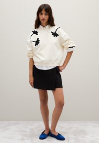 Felpa crema con applicazioni floreali nere, indossata su una camicia bianca con colletto. Abbinata a una gonna nera e scarpe slip-on blu.