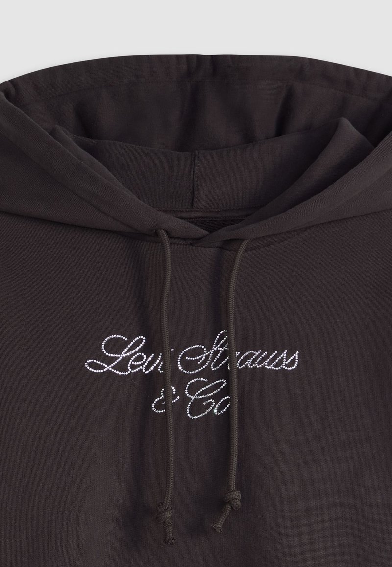 Sweat à capuche marron foncé en tissu doux, orné d'un logo en sequins de couleurs claires. Cordon de serrage avec un finis noué au niveau du col.