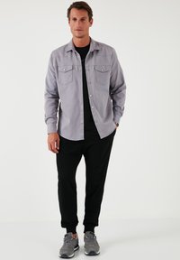 Cămașă butonată de culoare gri deschis, cu două buzunare la piept, purtată peste un tricou negru, asociată cu pantaloni sport negri și adidași gri.