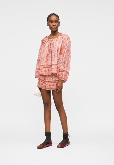 Femme aux cheveux tressés portant un haut à manches longues rose à motif cachemire et un short assorti, tenant un petit sac blanc, avec des chaussettes foncées et des chaussures rouges.