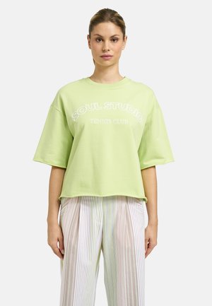 Woman wearing a light green oversized T-shirt with "SOUL STUDIO TENNIS CLUB" text and loose striped pastel trousers against a plain background.