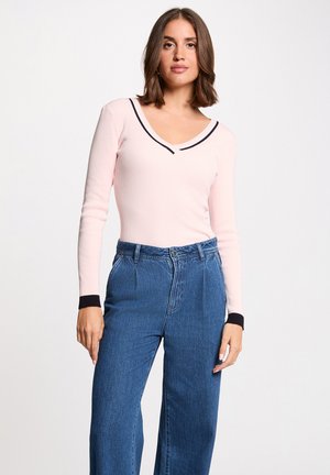 Mujer que lleva una camiseta de canalé de manga larga color rosa claro con ribete negro en el escote y los puños, combinada con vaqueros azules de talle alto, de pie contra un fondo blanco.