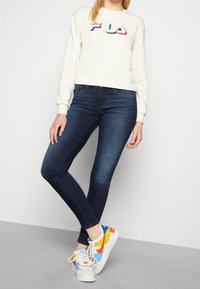 Persona che indossa una felpa bianca FILA, jeans skinny blu scuro e sneakers Nike multicolori, in piedi con le gambe incrociate.