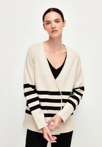 Beige cardigan med svarta horisontella ränder, v-ringning, dubbla knappar och sidofickor, med en mjuk textur.