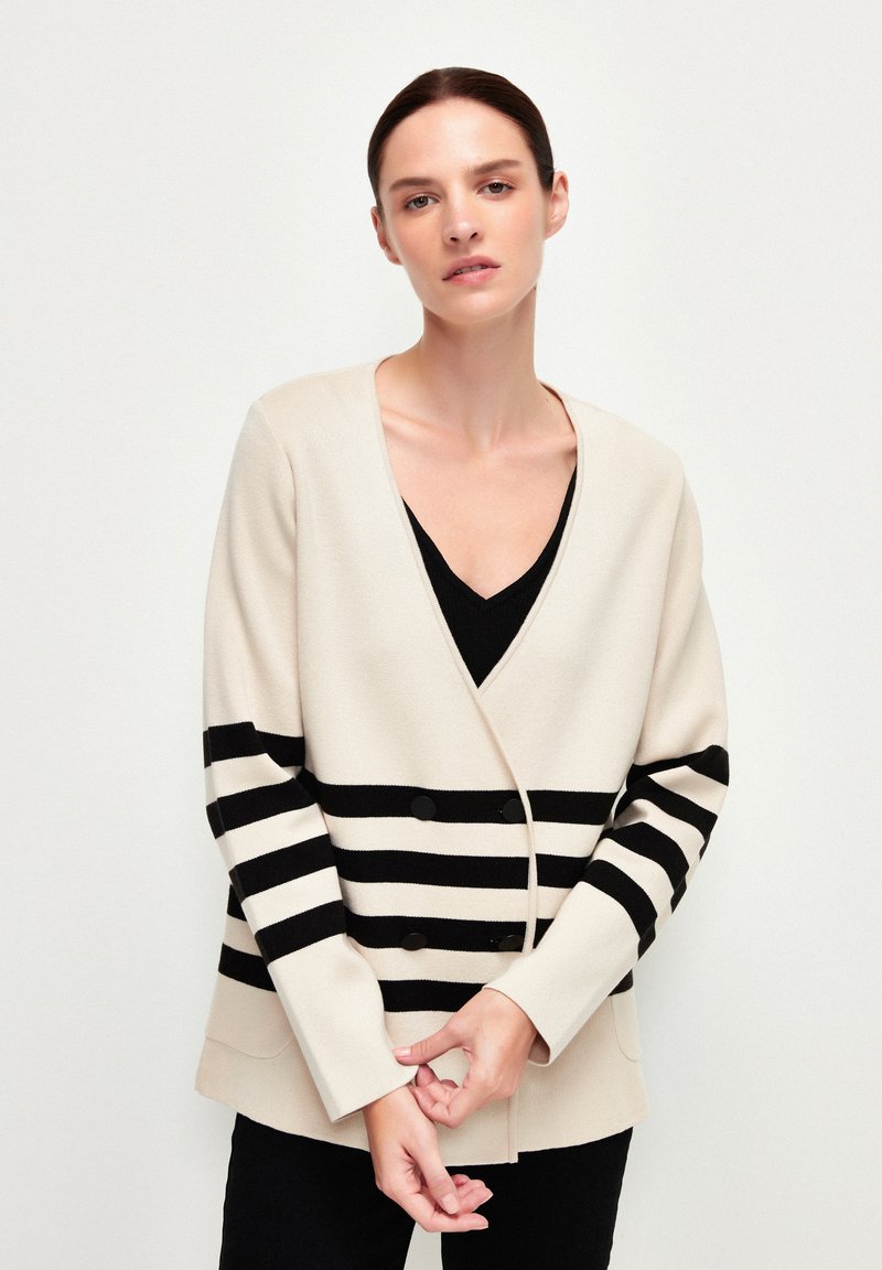 Beige cardigan med svarta horisontella ränder, v-ringning, dubbla knappar och sidofickor, med en mjuk textur.