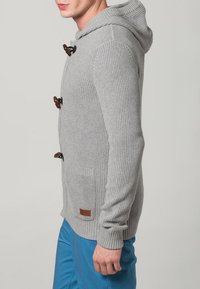 Šedá pletená mikina s dřevěnými zapínáními na knoflíky, žebrovanou texturou, bočními kapsami a malou hnědou koženou štítkem na spodní straně.