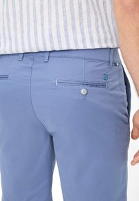 Pantaloni azzurri chiari caratterizzati da una trama liscia, una tasca posteriore con bottone e dettagli di cucitura sottili. Gli accenti includono un piccolo etichetta blu sul lato.