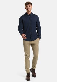 Lächelnder Mann, der vorwärts geht, trägt ein marineblaues Hemd mit Knöpfen, beigefarbene Hose und braune Schnürstiefel, mit einer Hand in der Tasche und einer ausgestreckten Hand.