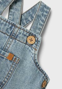 Denim salopete, kipe iz svetlo modrega blaga z zbledelim vzorcem, lesenimi gumbi in kontrastnimi oranžnimi šivi.