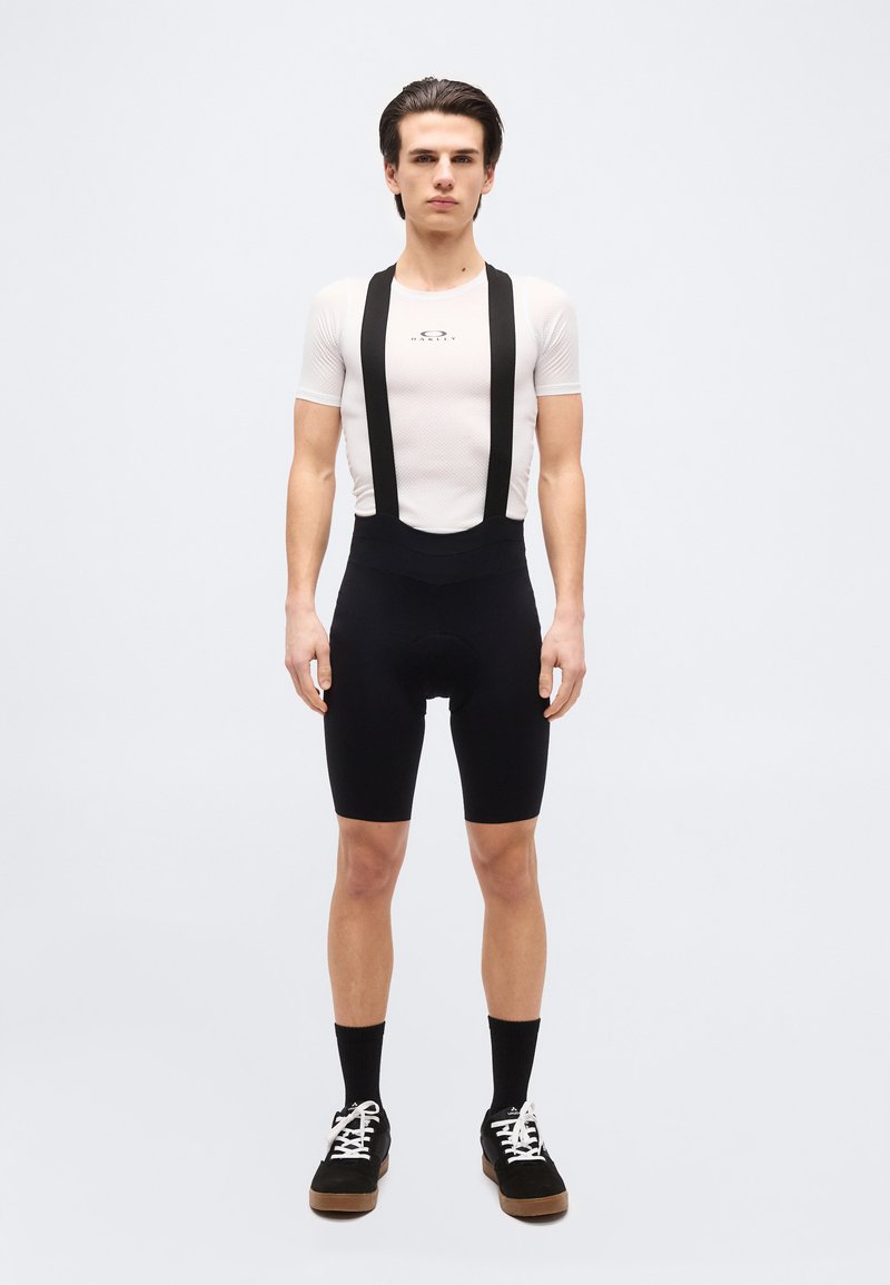 Nuori mies seisoo, pukeutuneena valkoiseen lyhythihaiseen pyöräilypohjakerrokseen ja mustiin pyöräilyhousuihin mustien sukkien ja mustien lenkkareiden kanssa.