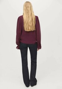 Blouse à manches longues, bordeaux avec des épaules bouffantes, associée à un jean en denim foncé évasé. Les cheveux sont longs et ondulés, portés détachés.
