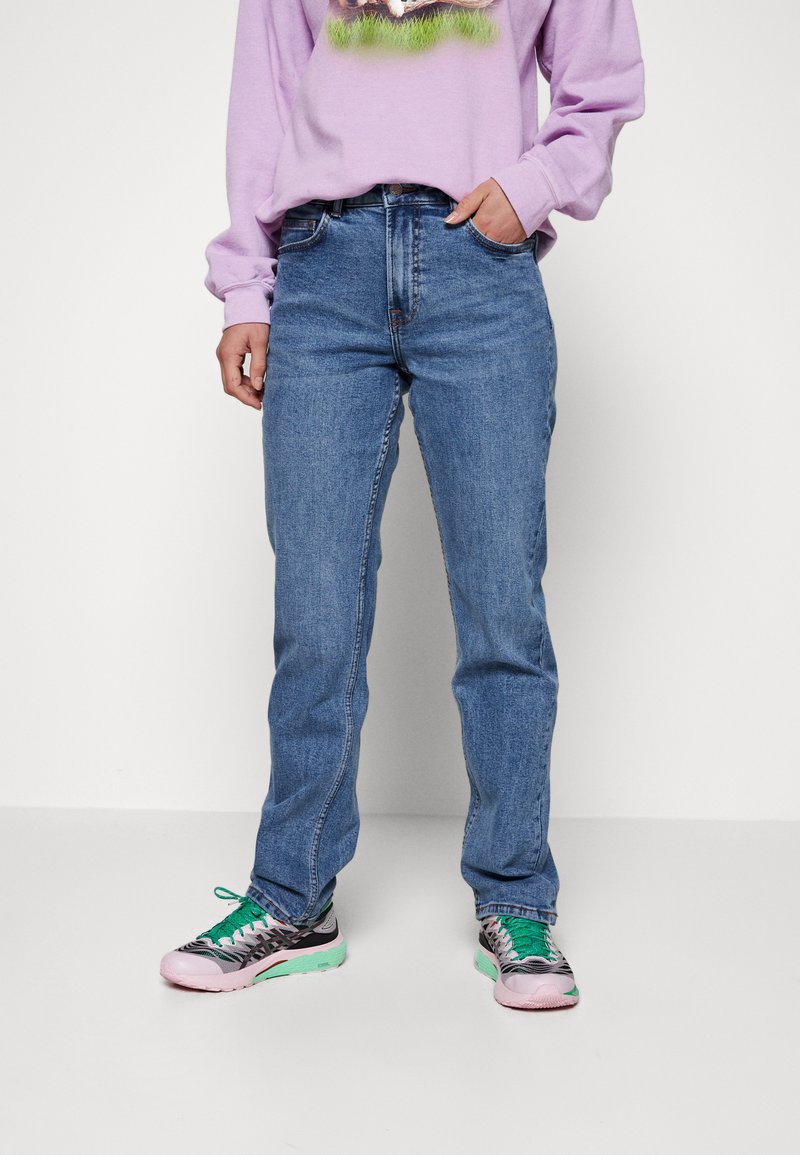 Vaqueros de mezclilla azules con corte recto, que presentan un diseño clásico de cinco bolsillos y costuras sutiles. Combinados con zapatillas de varios colores.