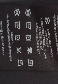 Etichetta di cura su tessuto nero con simboli di cura. Include istruzioni per il lavaggio, la stiratura e l'asciugatura in bianco, con menzione di un marchio ecologico.