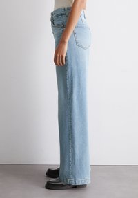 Světle modré džíny s vysokým pasem a rovnými nohavicemi, vyrobené z denimu. Mají zadní kapsy a jsou stylizovány s černými platformovými botami.