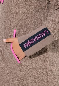 Giacca di pile marrone con una texture morbida, dotata di dettagli rosa, tasche frontali e un polsino scuro con la scritta "LAURASON" in lettering rosa.