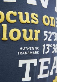 Marineblauer Stoff mit erhabenen gelben und weißen Texten, darunter "focus on your", Koordinaten und "AUTHENTIC TRADEMARK" in Großbuchstaben.