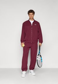 Jachetă și pantaloni de trening de culoare bordo, fabricați dintr-un material ușor. Caracteristici: guler înalt, manșete elastice și închidere cu fermoar. Ține o rachetă de tenis și o minge.