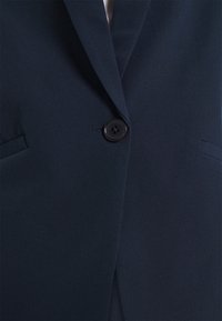 Enkeltknappet mørkeblå blazer med hakkede revers og to horisontale forlommer vist i nærbillede.