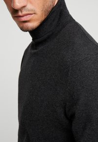 Pull à col roulé gris foncé en tissu tricoté doux, présentant une coupe ajustée et un col côtelé, porté par un modèle vu de profil.