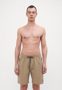 Shorts de bain beiges avec une taille élastique, cordon noir, poches zippées sur les côtés et un écusson logo sur la jambe inférieure. Texture de tissu lisse.