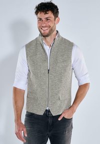 Graue, strukturierte Weste mit einem Frontreißverschluss und zwei Taschen. Über einem weißen Hemd getragen, hat die Weste einen offenen Kragen und einen taillierten Schnitt.