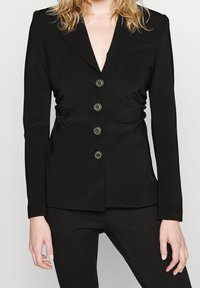 Černé na míru šité sako s hlubokým výstřihem do V, pětimístným zapínáním na knoflíky s kovovými detaily, vypolstrovanými rameny a přiléhavým střihem.