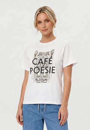 Femme blonde portant un T-shirt blanc avec l'inscription « Café Poésie » et un motif de porte de Paris, associée à un jean bleu, debout devant un fond uni.
