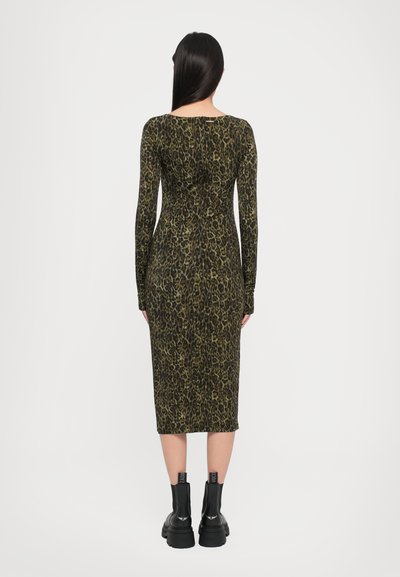 Robe ajustée à manches longues en imprimé léopard vert et noir, arrivant au milieu du mollet. Portée avec des bottines noires à talons épais et à languettes.