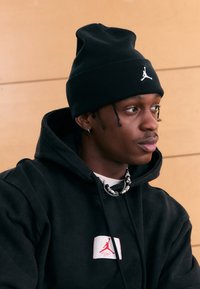 Svart lue med en hvit logo, svart hettegenser med en rød og hvit lapp, flerlags halskjeder, mot en lys trebakgrunn.