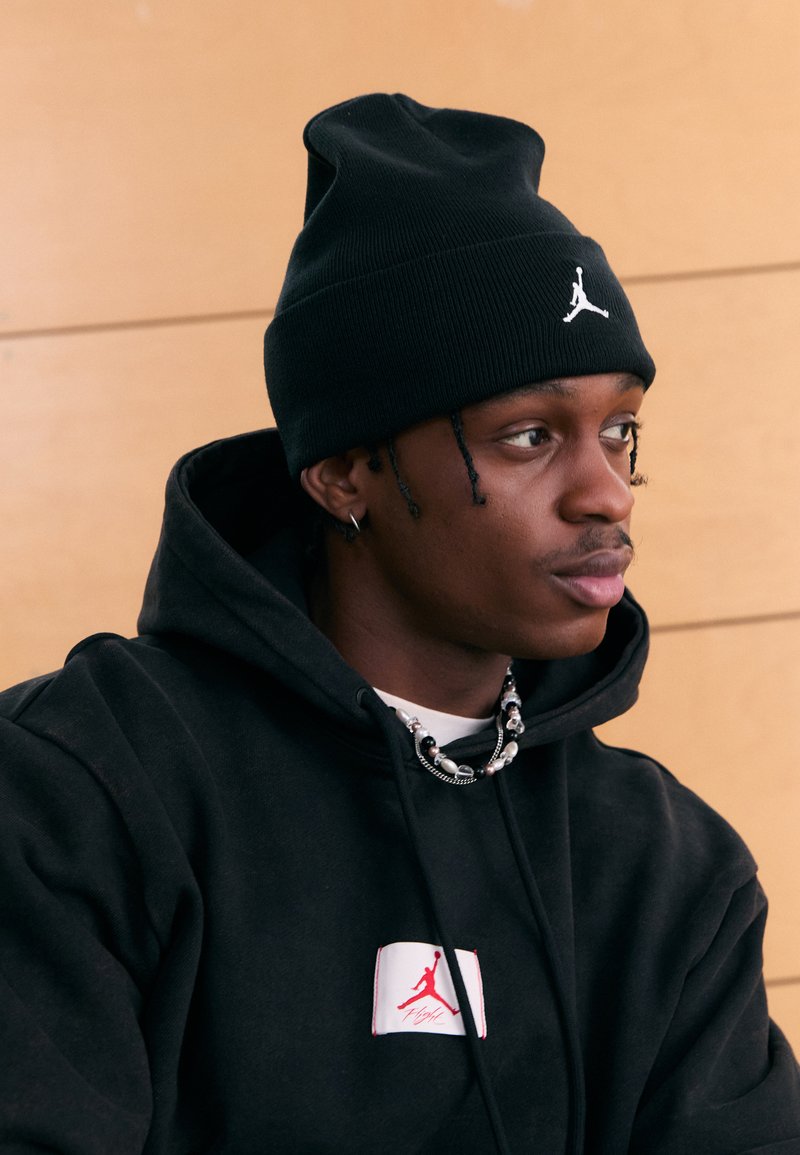 Svart lue med en hvit logo, svart hettegenser med en rød og hvit lapp, flerlags halskjeder, mot en lys trebakgrunn.