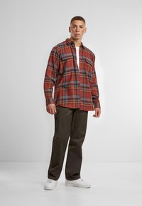Red and blue plaid flannel shirt with long sleeves and two front pockets, paired with dark green pants and white sneakers.