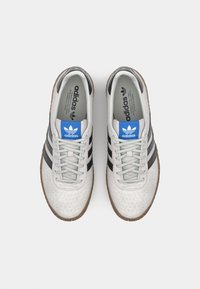 Grå perforerede sneakers med tre sorte striber, teksturerede grå snørebånd og et blåt logo-patch på tungen. Brun gummisål.
