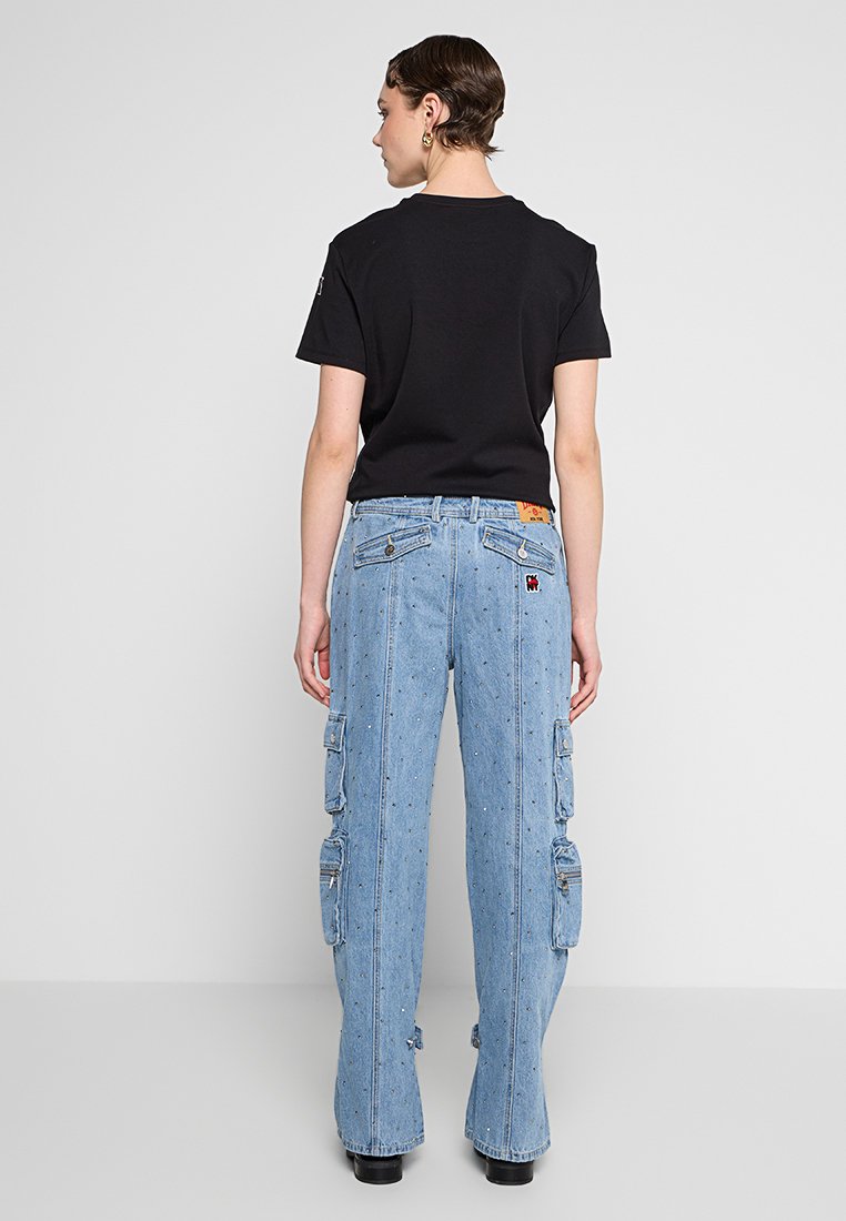 Frau, die mit dem Rücken zur Kamera steht, trägt ein schwarzes T-Shirt und hellblaue Denim-Cargo-Jeans mit kleinen Verzierungen sowie schwarze Schuhe.