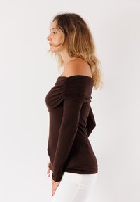 Top marrone scollato con dettagli arricciati sul petto e maniche lunghe aderenti. Tessuto liscio, design attillato, indossato con pantaloni bianchi.