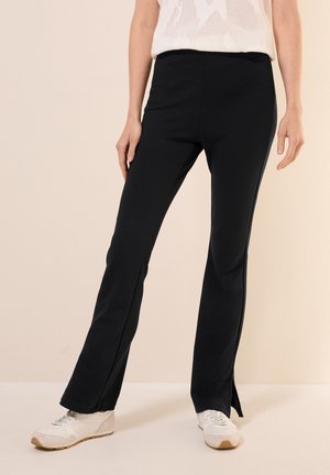 Schwarze dehnbare Leggings mit weit auslaufender Form und seitlichen Schlitz am Saum, kombiniert mit weißen Sportschuhen. Strukturiertes weißes Oberteil teilweise sichtbar.